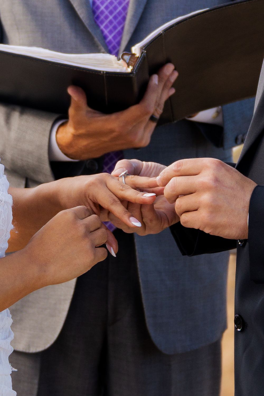 Groom placing a wedding ring on brides finger during an outdoor ceremony officiated by a minister.