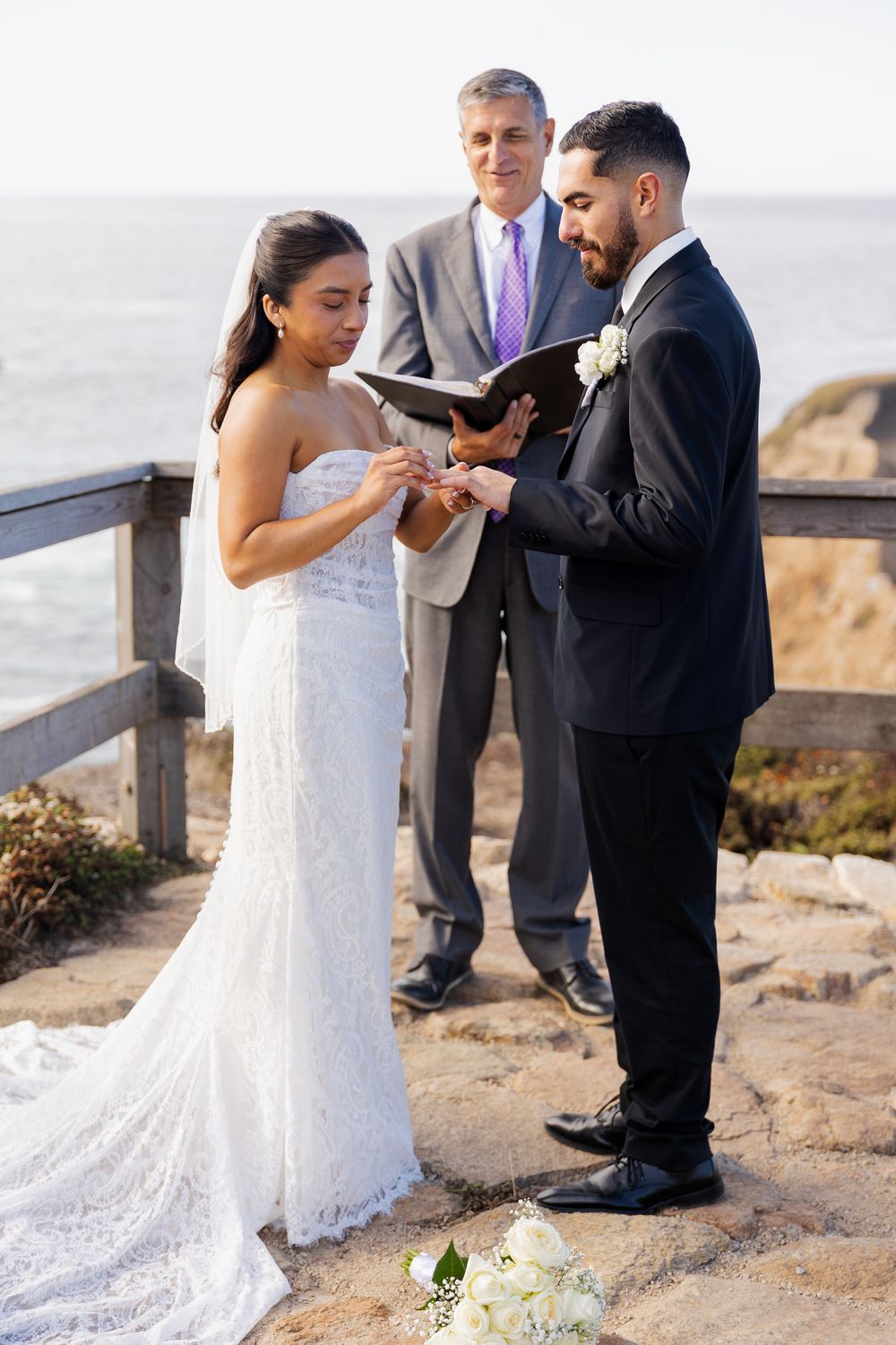 Bride places wedding ring on groom's finger during outdoor coastal ceremony with officiant.