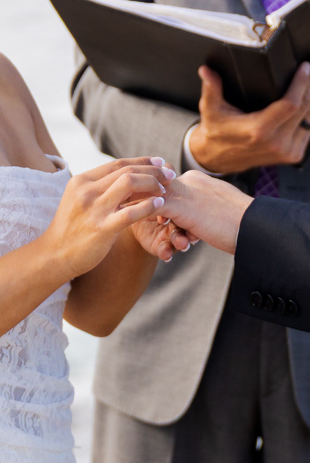 Bride placing a wedding ring on groom's finger during ceremony, with officiant holding book in background.