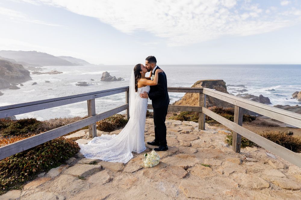 Newlyweds share first kiss on rocky ocean overlook with stunning Big Sur coastline backdrop.