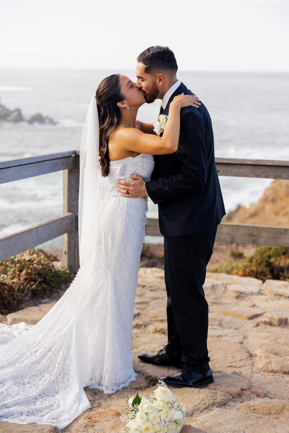 Bride and groom share a romantic kiss on a coastal cliff, with ocean views, in wedding attire.
