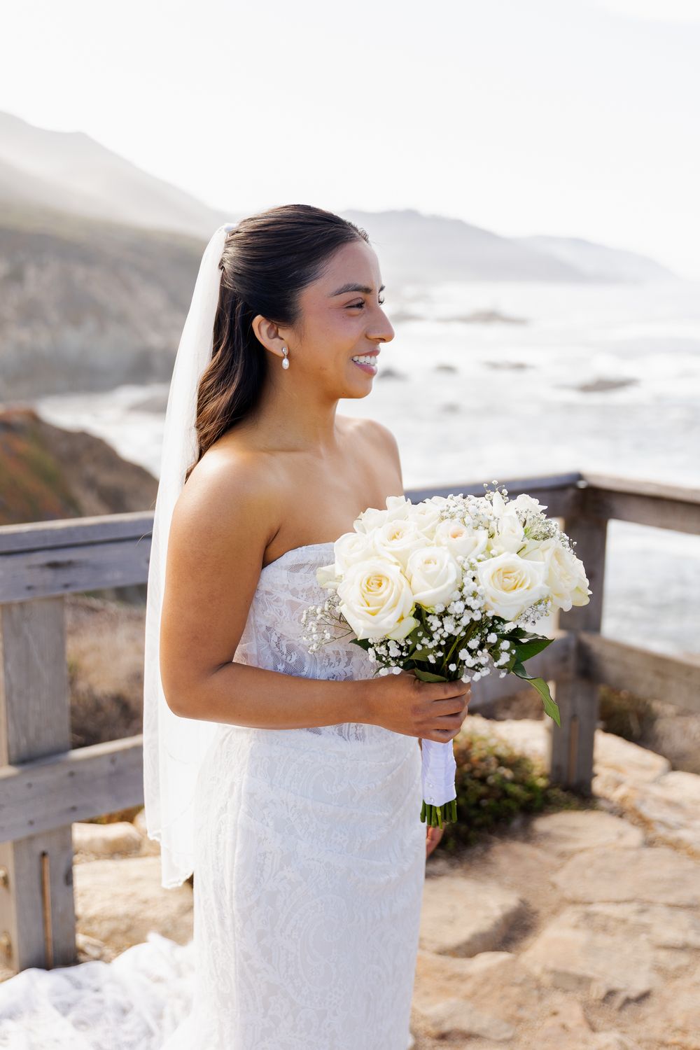 Smiling bride in strapless white lace gown holds white rose bouquet on scenic coastal overlook.