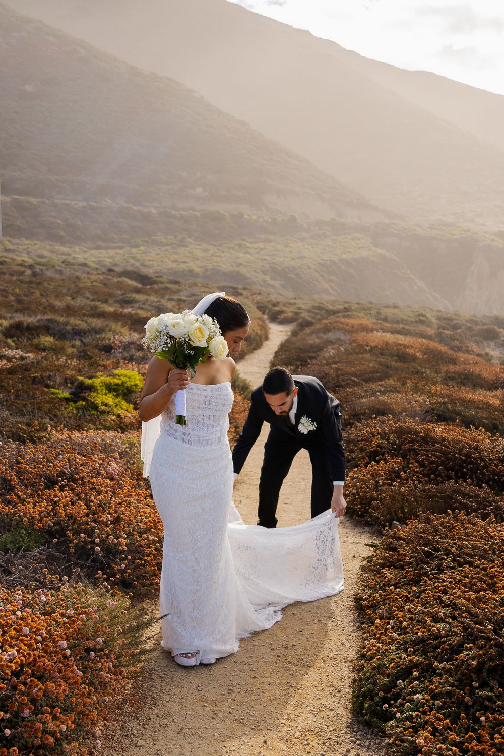 Groom adjusting bride's white lace wedding dress train on a scenic coastal trail surrounded by golden shrubs.