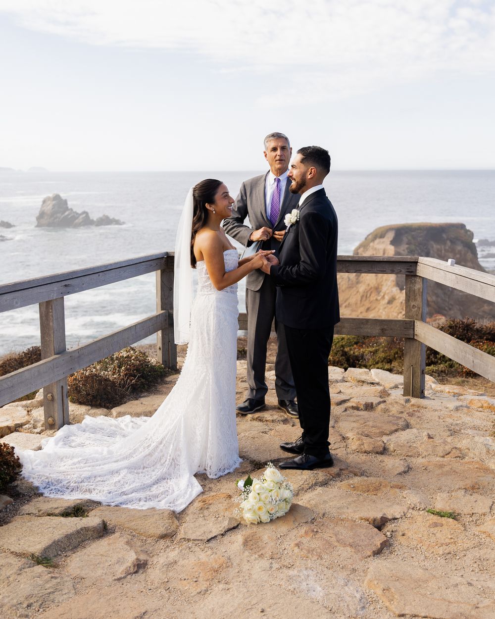 Couple exchanging vows at a cliffside ocean wedding ceremony with an officiant, bride in white lace gown with veil.