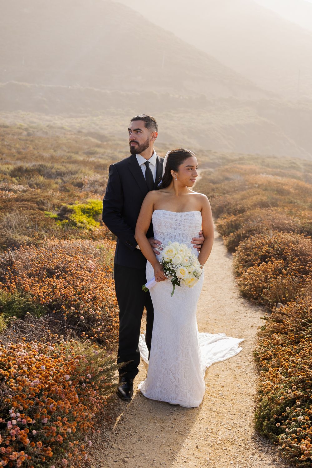 Bride and groom pose on a scenic coastal path surrounded by wildflowers during golden hour wedding photography.