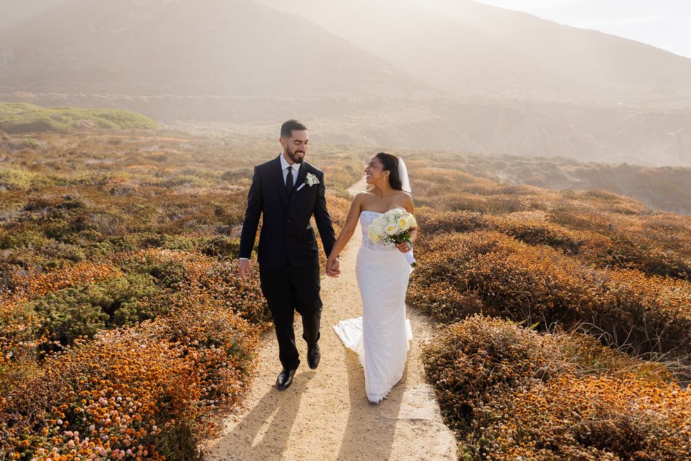 Bride and groom walk hand-in-hand through coastal wildflowers at golden hour wedding in Big Sur.