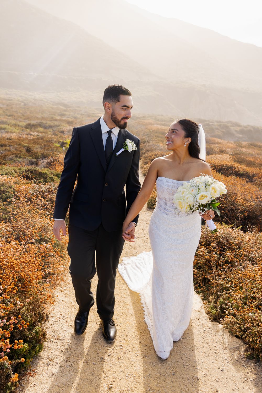 Newlyweds walk hand in hand along a sunlit coastal trail, bride holding white bouquet in wedding attire.