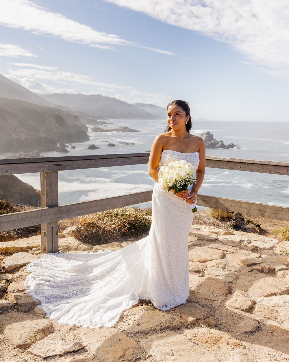 Bride in white lace gown holding bouquet on coastal overlook with ocean cliffs backdrop.