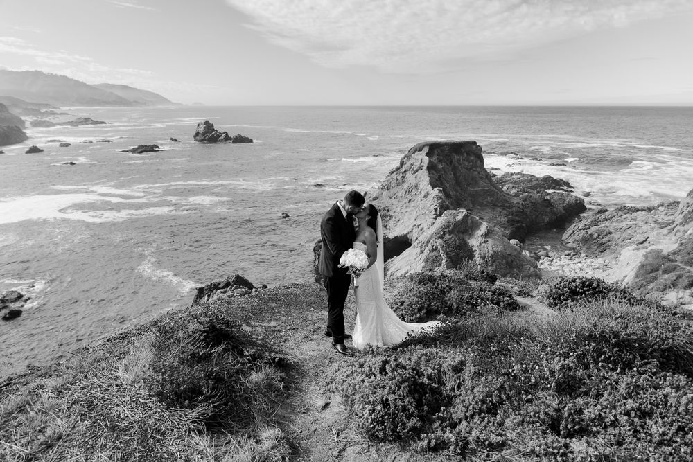 Bride and groom kissing on rocky coastal cliffs with ocean waves in black and white wedding photo.