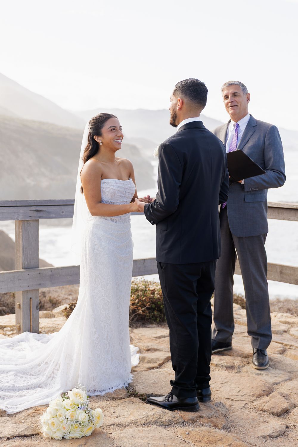 Bride and groom exchange vows at outdoor coastal wedding ceremony with officiant holding book, ocean cliffs in background.