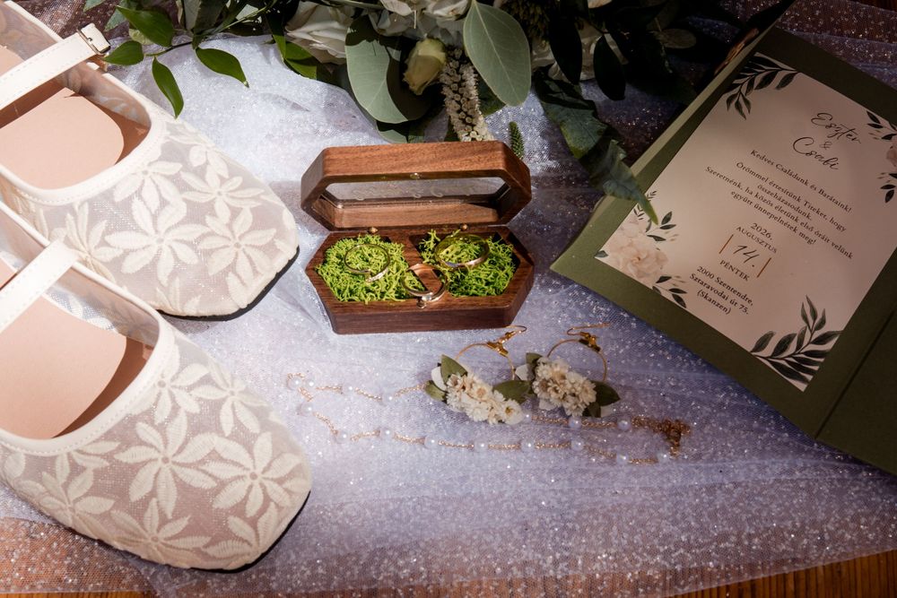 Bridal flat shoes, wooden ring box with moss and wedding rings, floral invitation on glittery surface.