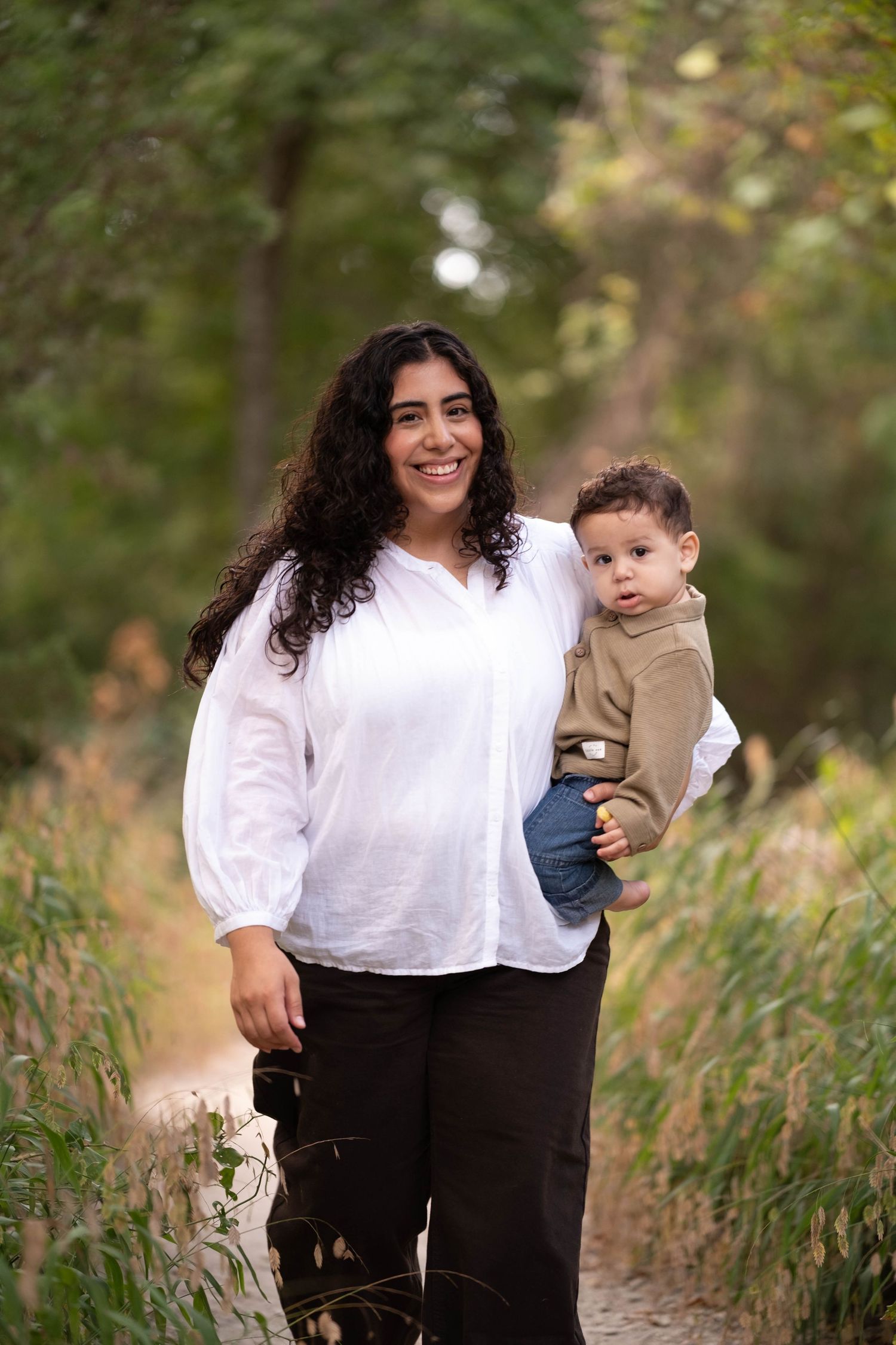 A smiling woman with curly dark hair in a white blouse holds a toddler boy on a nature path surrounded by greenery.