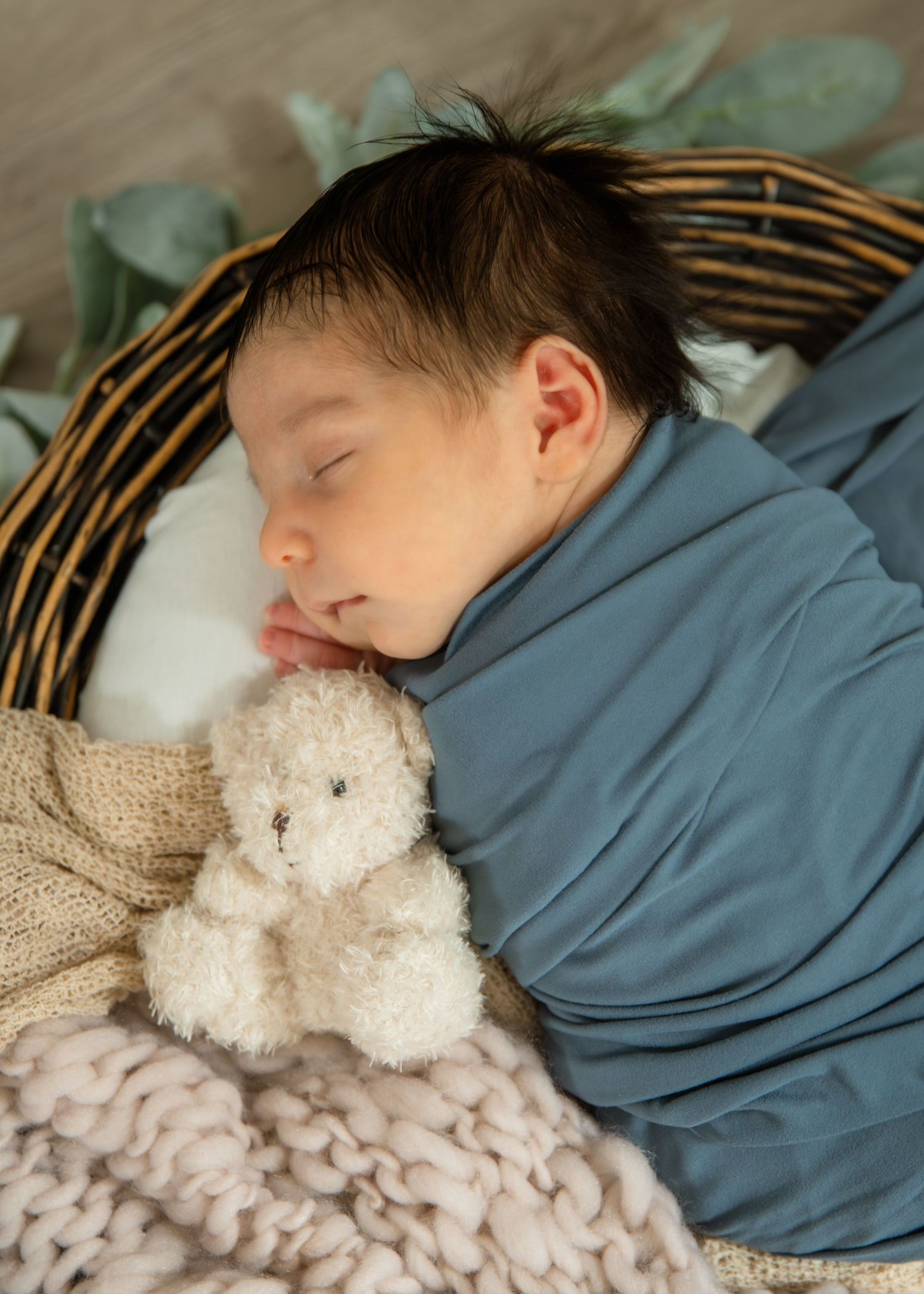 Newborn baby sleeping peacefully in a wicker basket, wrapped in a blue blanket, cuddling a small white teddy bear.