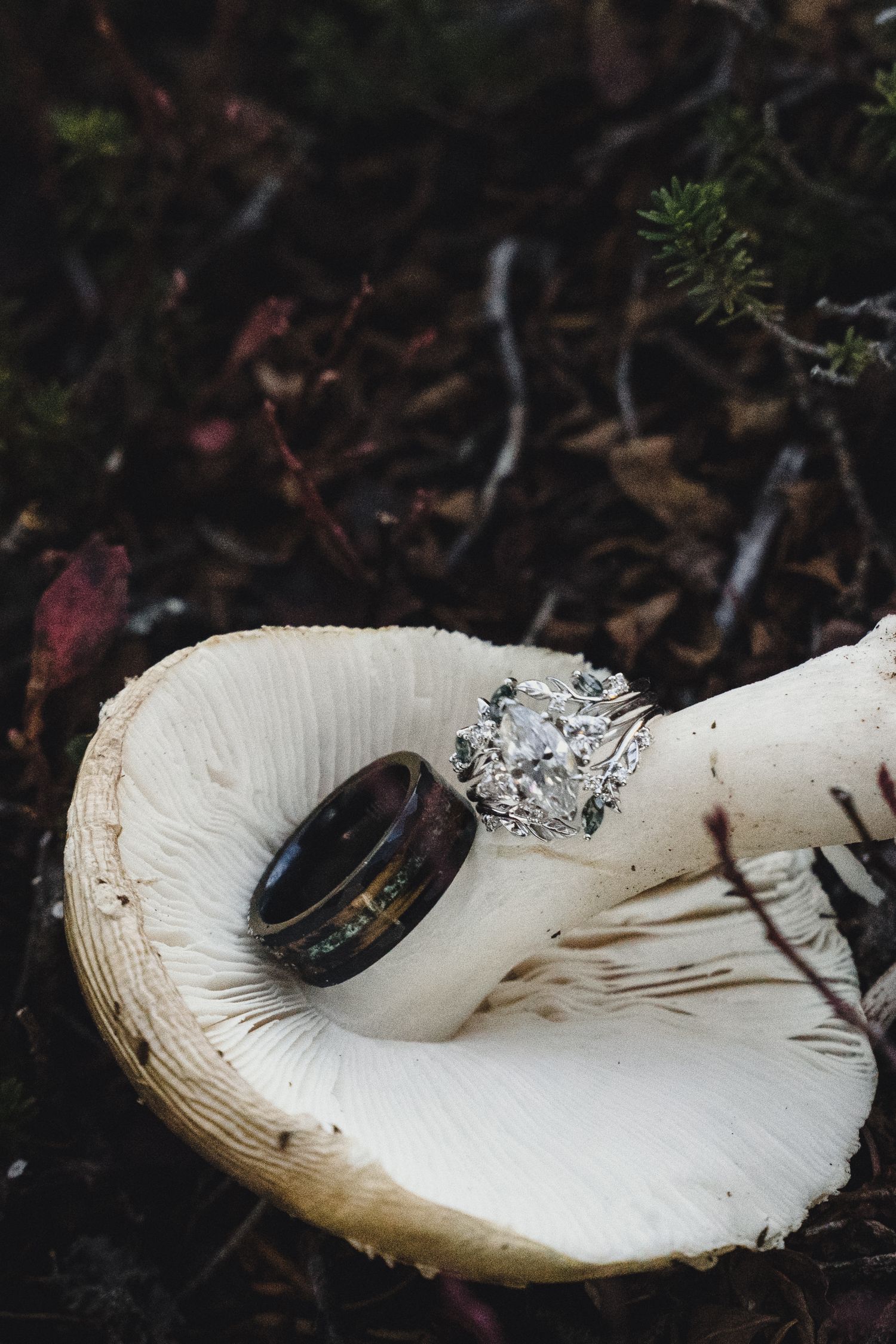 Diamond cluster engagement ring and dark wood wedding band resting on an overturned white mushroom among forest floor leaves and moss.