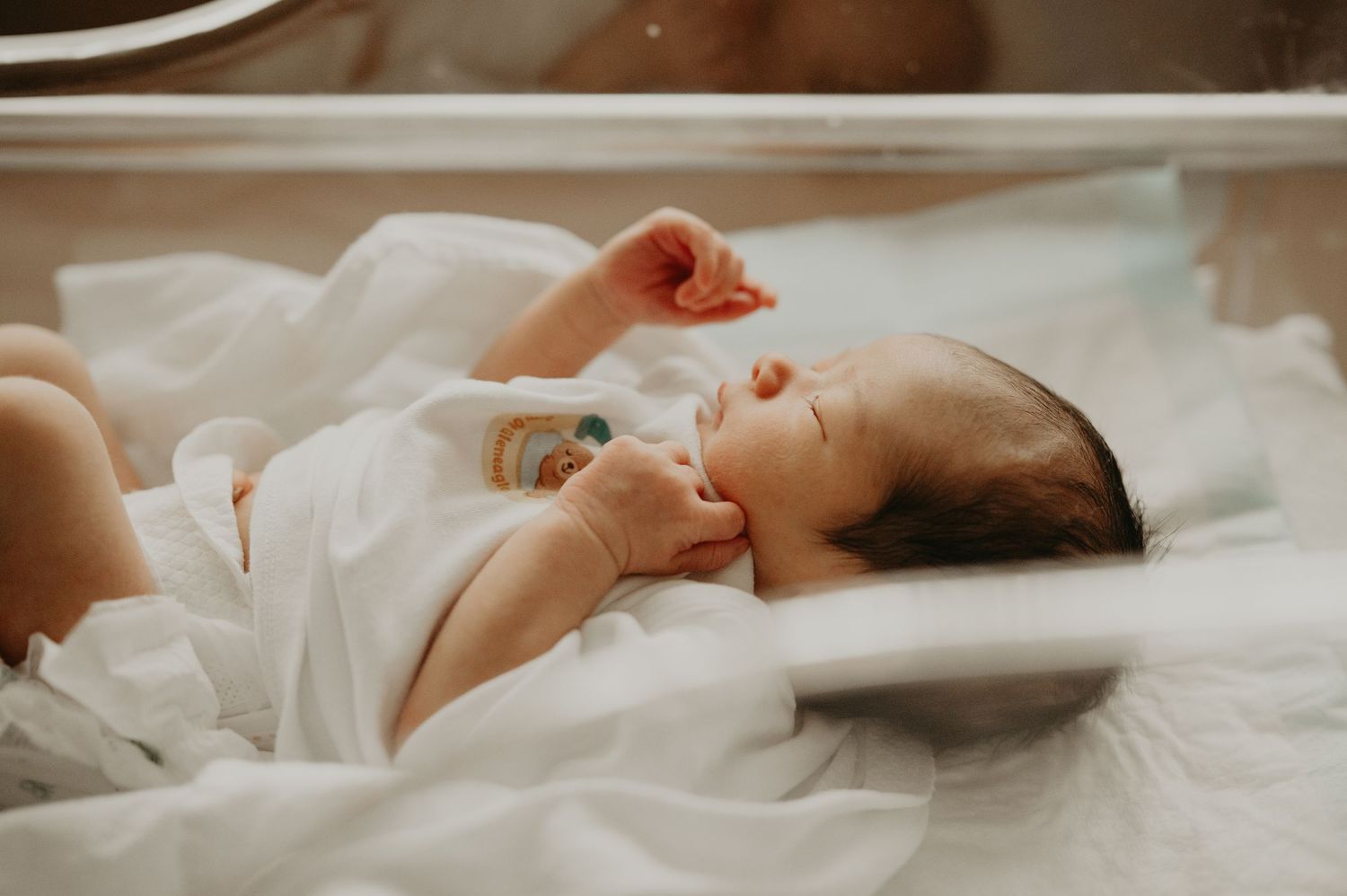 Sleeping newborn baby in a white onesie with hands near the face, resting peacefully in a clear hospital bassinet.