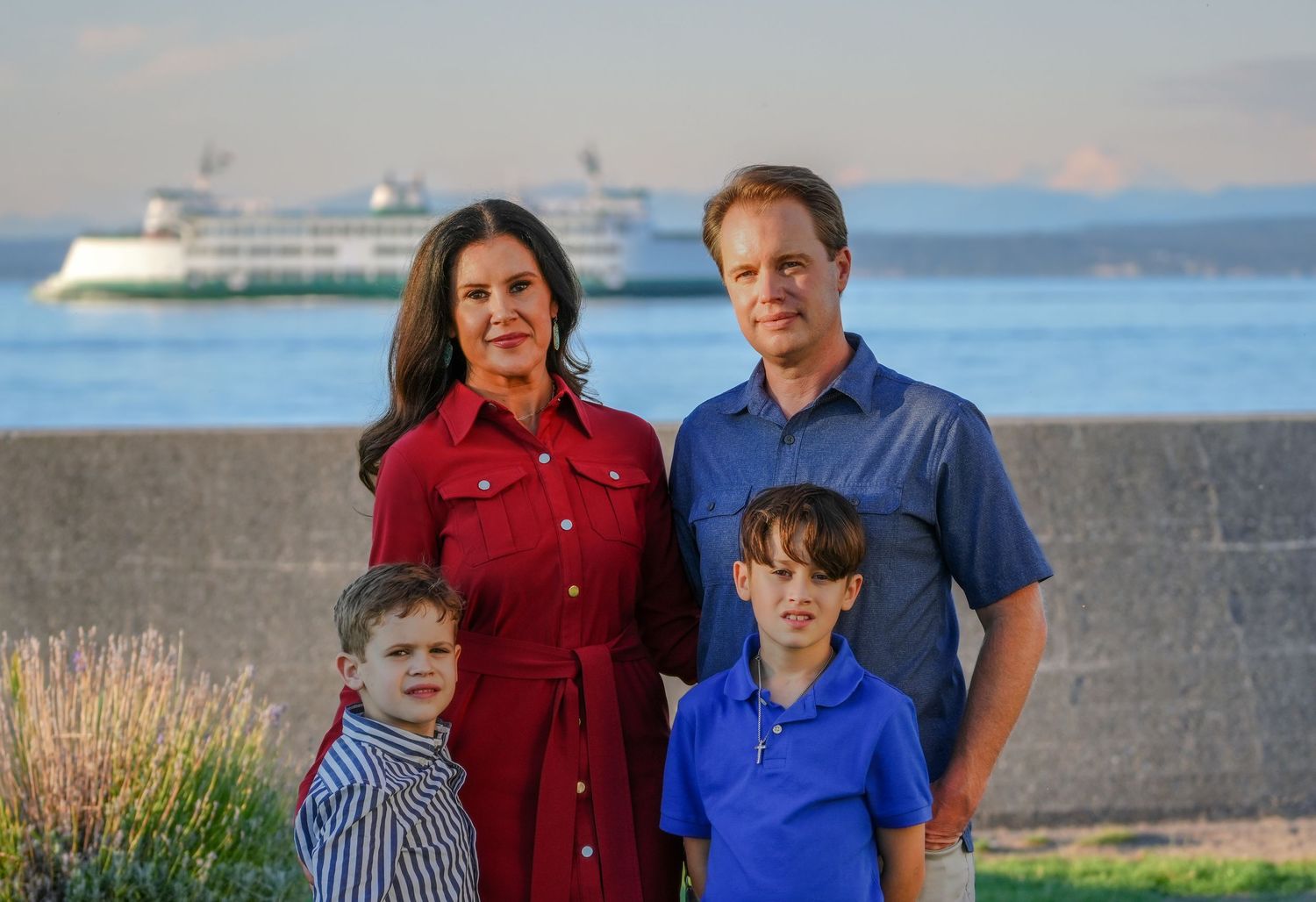 Family of four poses outdoors near waterfront with ferry boat and mountain in background on sunny day.