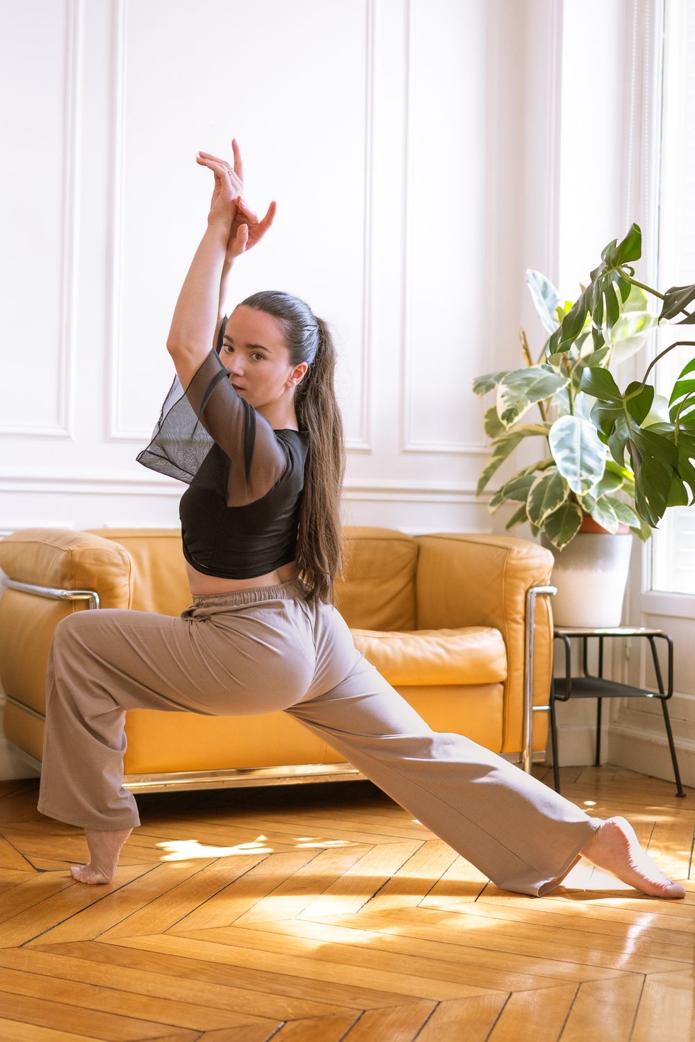 Danseuse en fente profonde, bras levé, queue-de-cheval, haut noir et pantalon taupe, près d'un canapé jaune