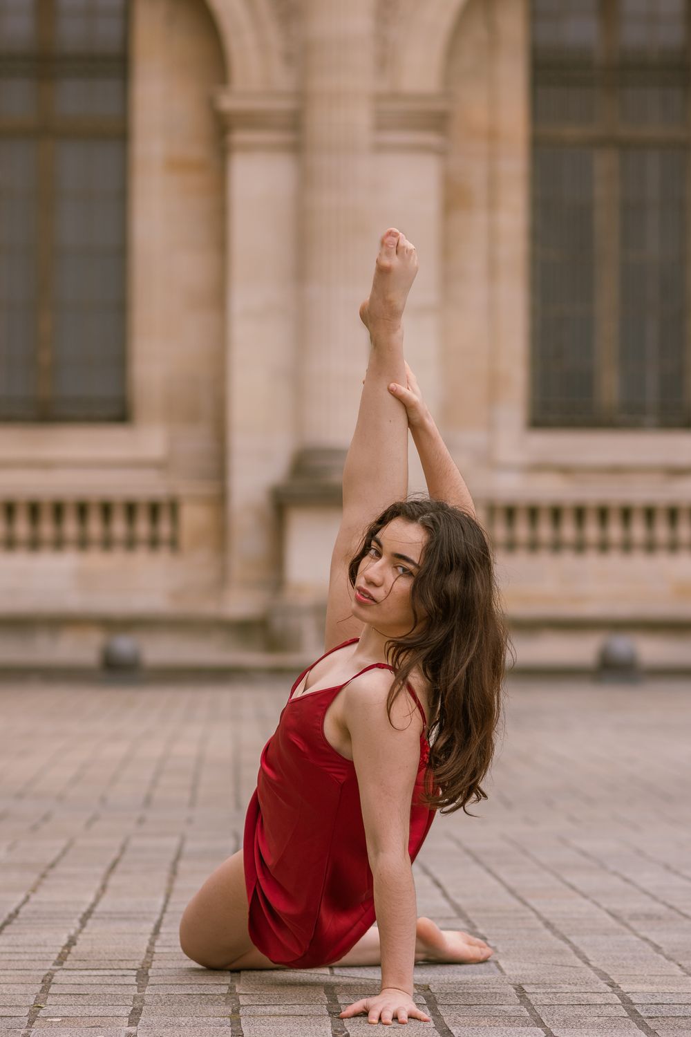 Danseuse aux longs cheveux bruns en robe satinée rouge, jambe tendue vers le ciel, sur un pavé devant un bâtiment de pierre