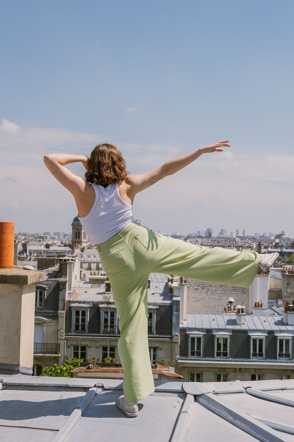Danseuse de dos, cheveux bruns ondulés, débardeur blanc et pantalon vert d'eau, jambe levée sur un toit zingué