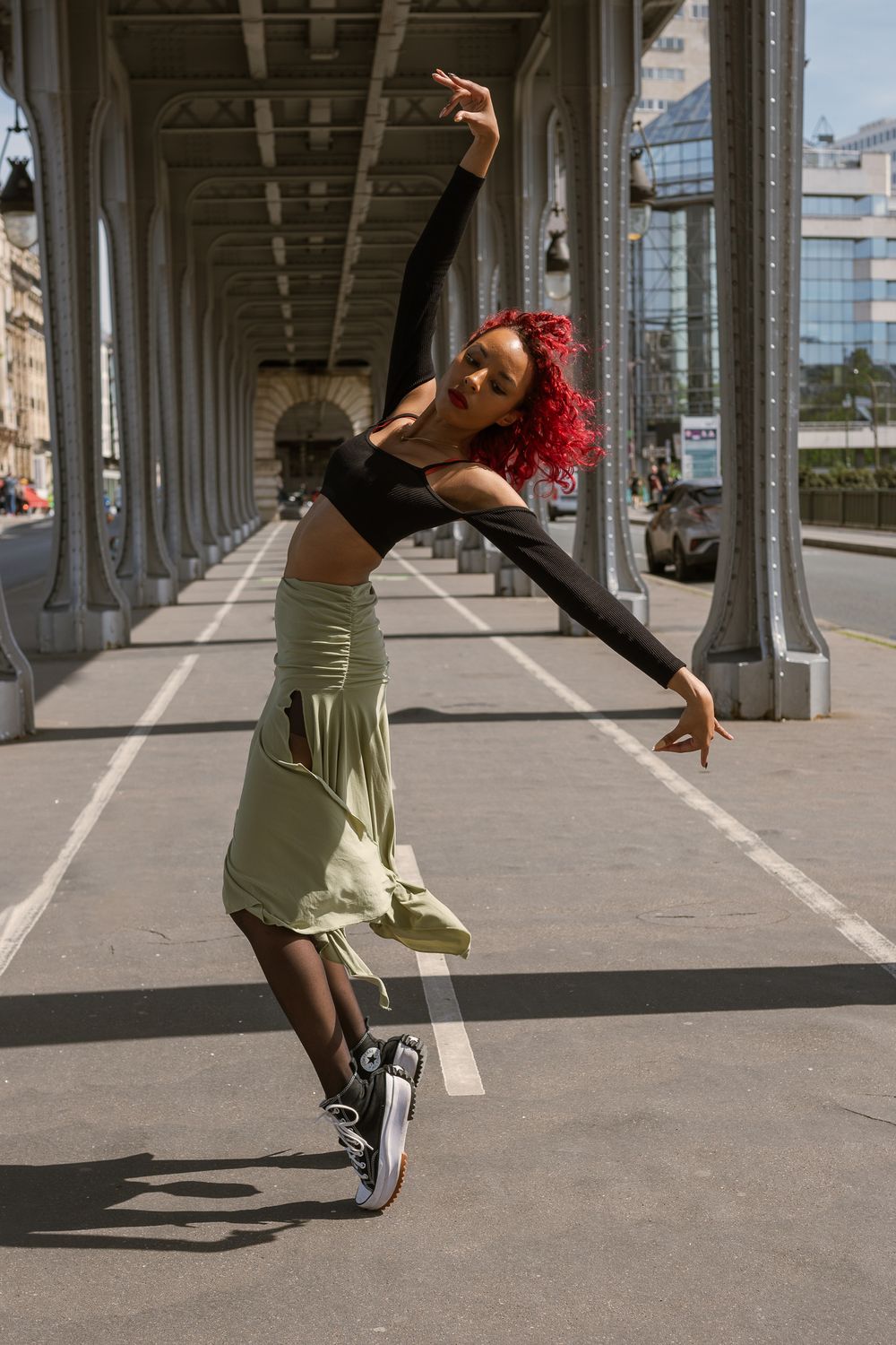 Danseuse aux cheveux bouclés rouges, haut noir et jupe vert sauge, bras levés en pleine pose sous un pont métallique