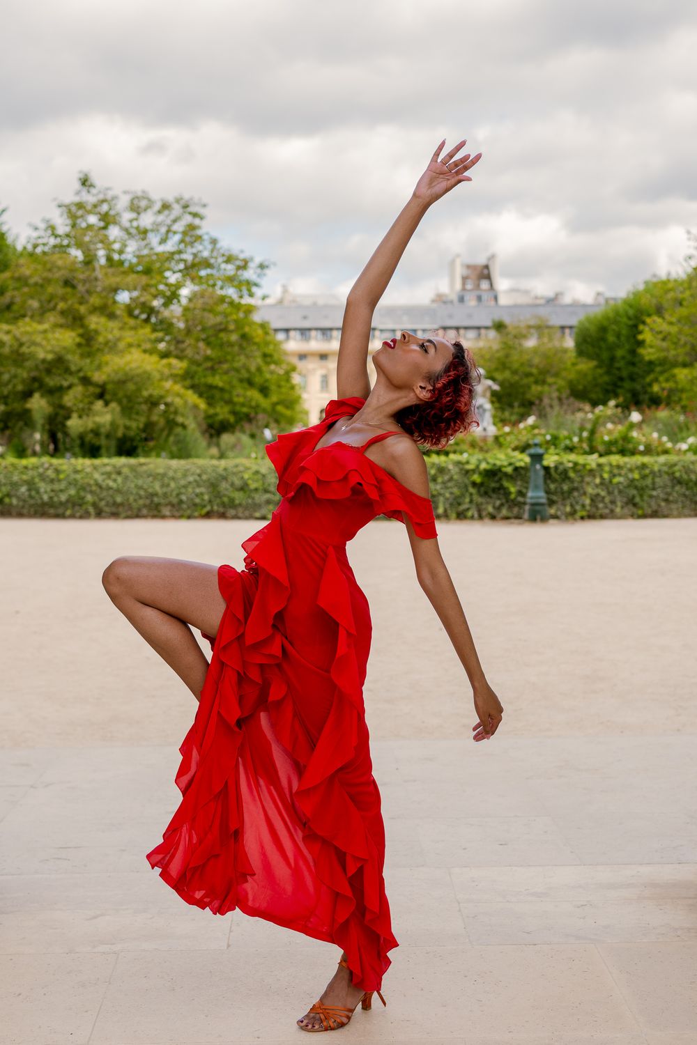 Danseuse aux cheveux courts bouclés roux, robe rouge à volants, bras levé et jambe pliée, dans un jardin