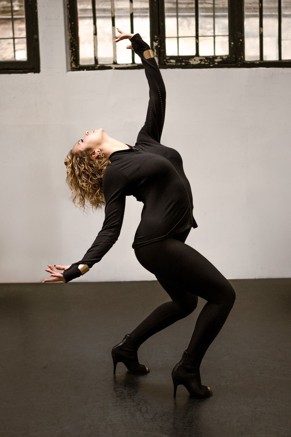 Danseuse aux cheveux bouclés blonds, tenue noire et talons, cambrée en arrière, bras levé, dans un studio aux grandes fenêtres