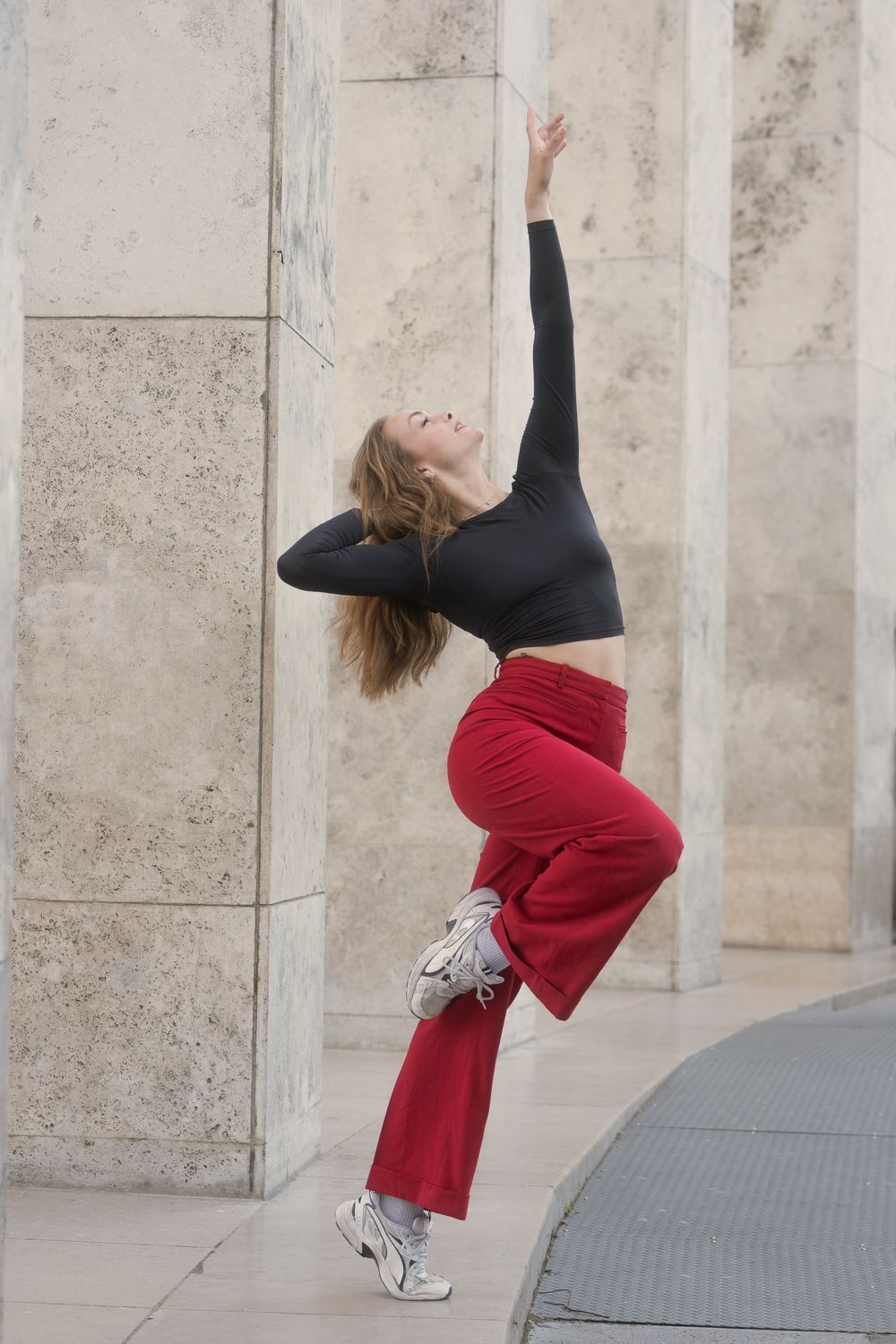 Danseuse aux longs cheveux châtains, haut noir et pantalon rouge, bras levé et genou plié, devant des piliers de pierre