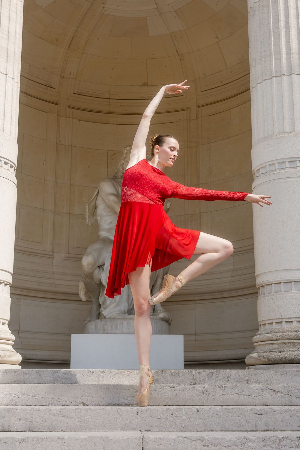 Danseuse en robe rouge à manches en dentelle, sur pointes, bras tendus, jambe repliée, devant une niche en pierre