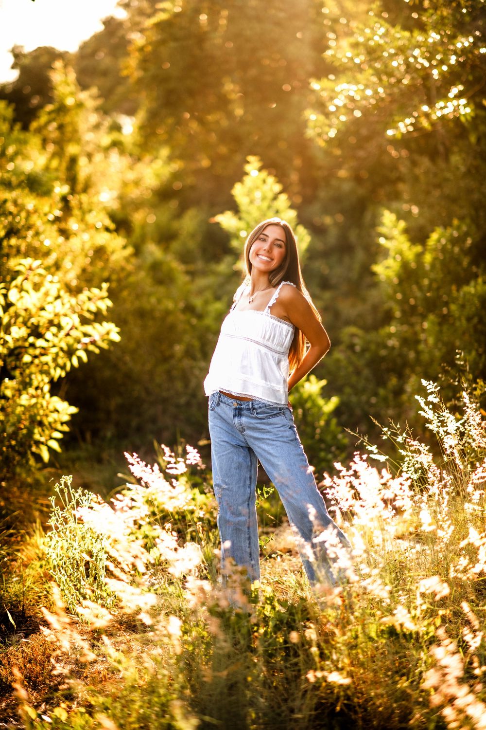 Smiling young woman in white top and jeans standing in sunlit nature with golden light filtering through green trees.