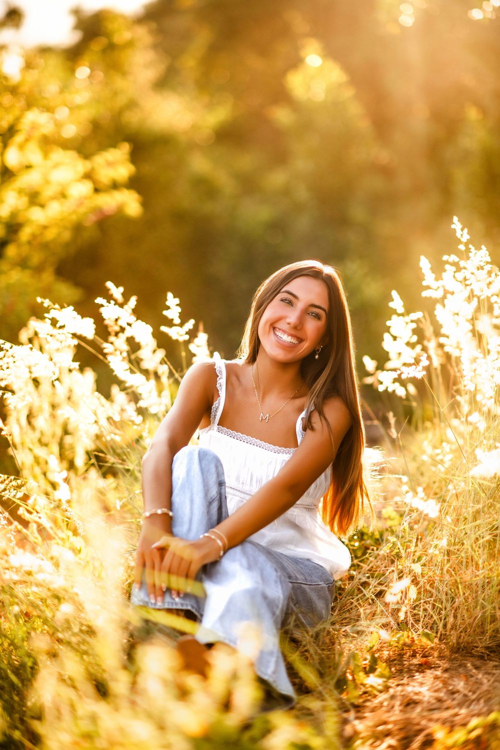 Young woman with long brown hair smiles while sitting in golden sunlit meadow wearing white tank top and blue jeans.