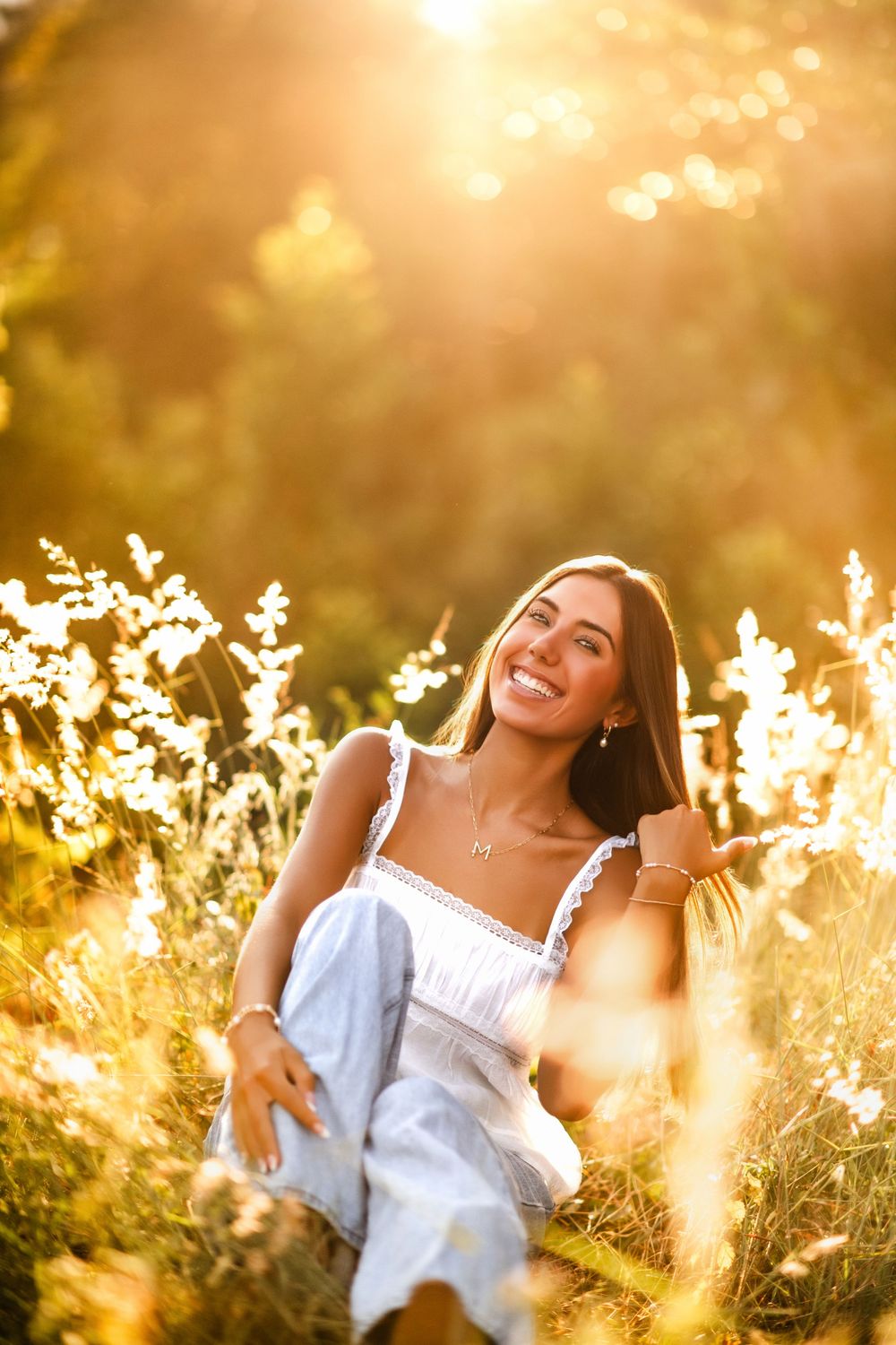 Young woman in white dress sitting in golden sunlit meadow with wildflowers, smiling warmly at sunset.