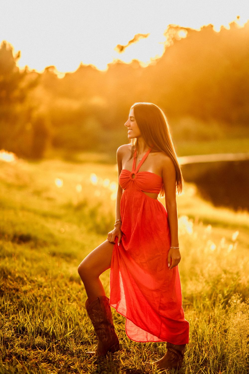 Woman in red halter maxi dress and cowboy boots standing in sunlit meadow at golden hour sunset.