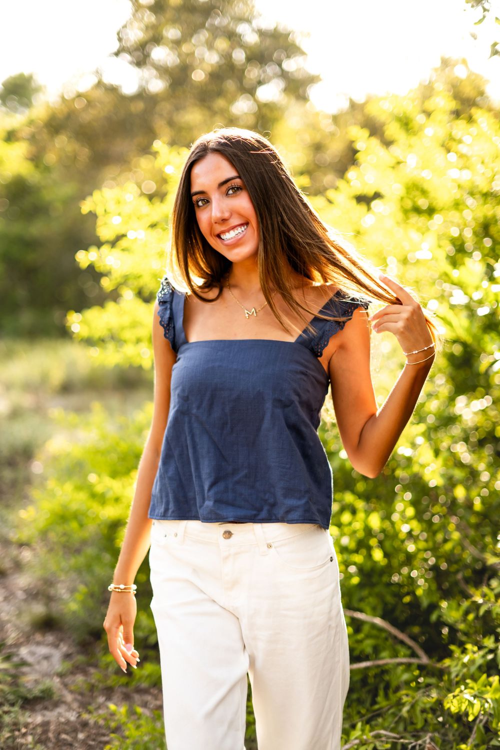 Young woman smiling outdoors in navy blue top and white pants, standing among lush green trees in golden sunlight.