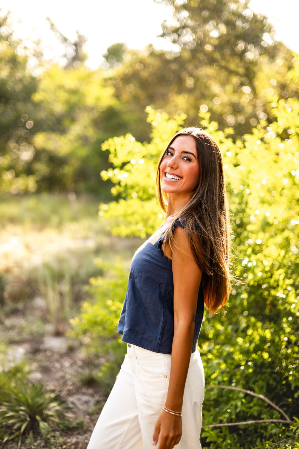 Smiling young woman in navy top and white pants standing outdoors in sunlit green nature setting.