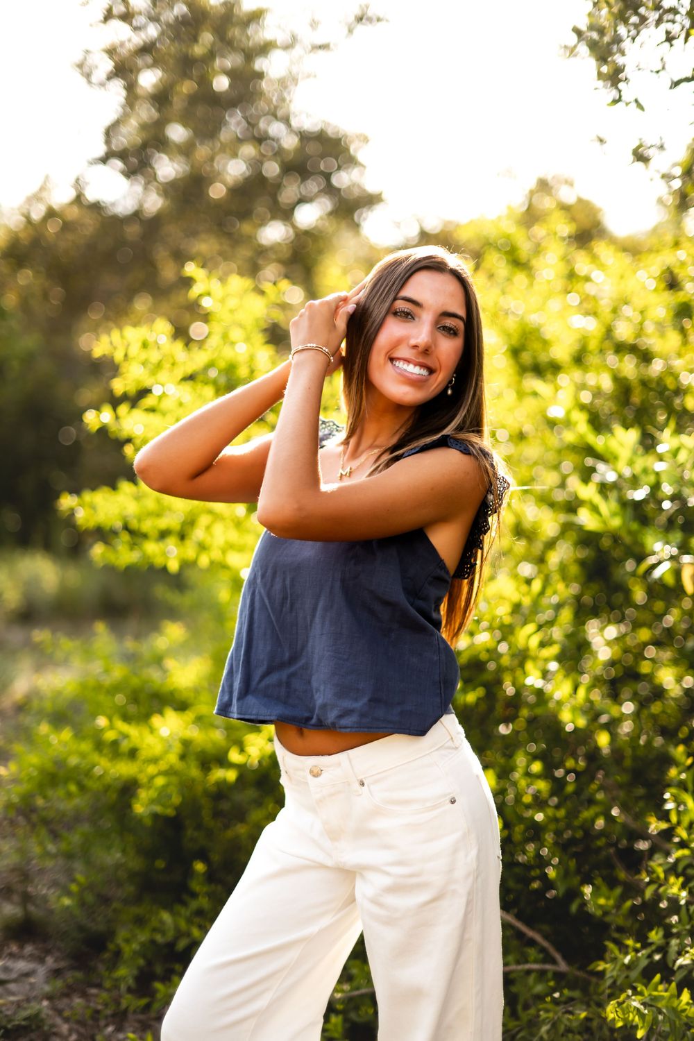 Young woman in navy crop top and white wide-leg pants smiling outdoors with lush green foliage background.