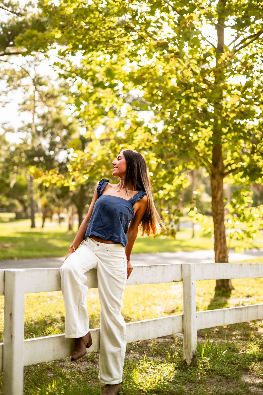 Young woman in blue top and white pants leaning on a white fence in a sunny green park.