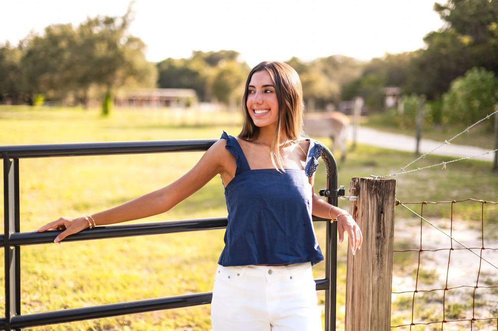 Young woman in navy top and white jeans leaning on a farm gate in a sunny rural field.
