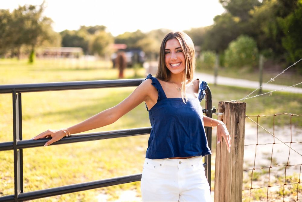 Young woman in blue top and white jeans smiling by a ranch fence in golden sunlight.