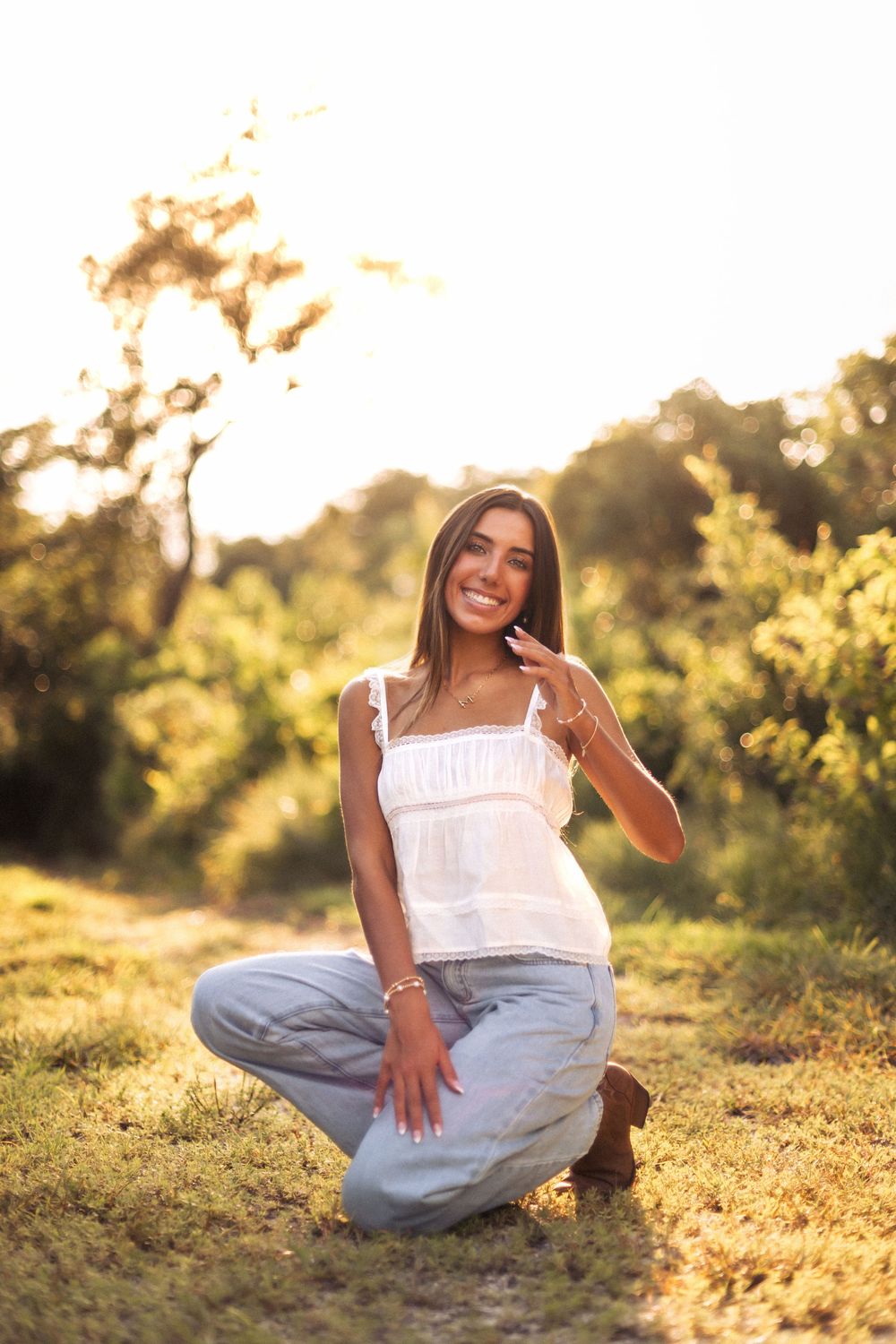 Young woman in white top and jeans crouching on grass in golden sunset light, smiling outdoors.