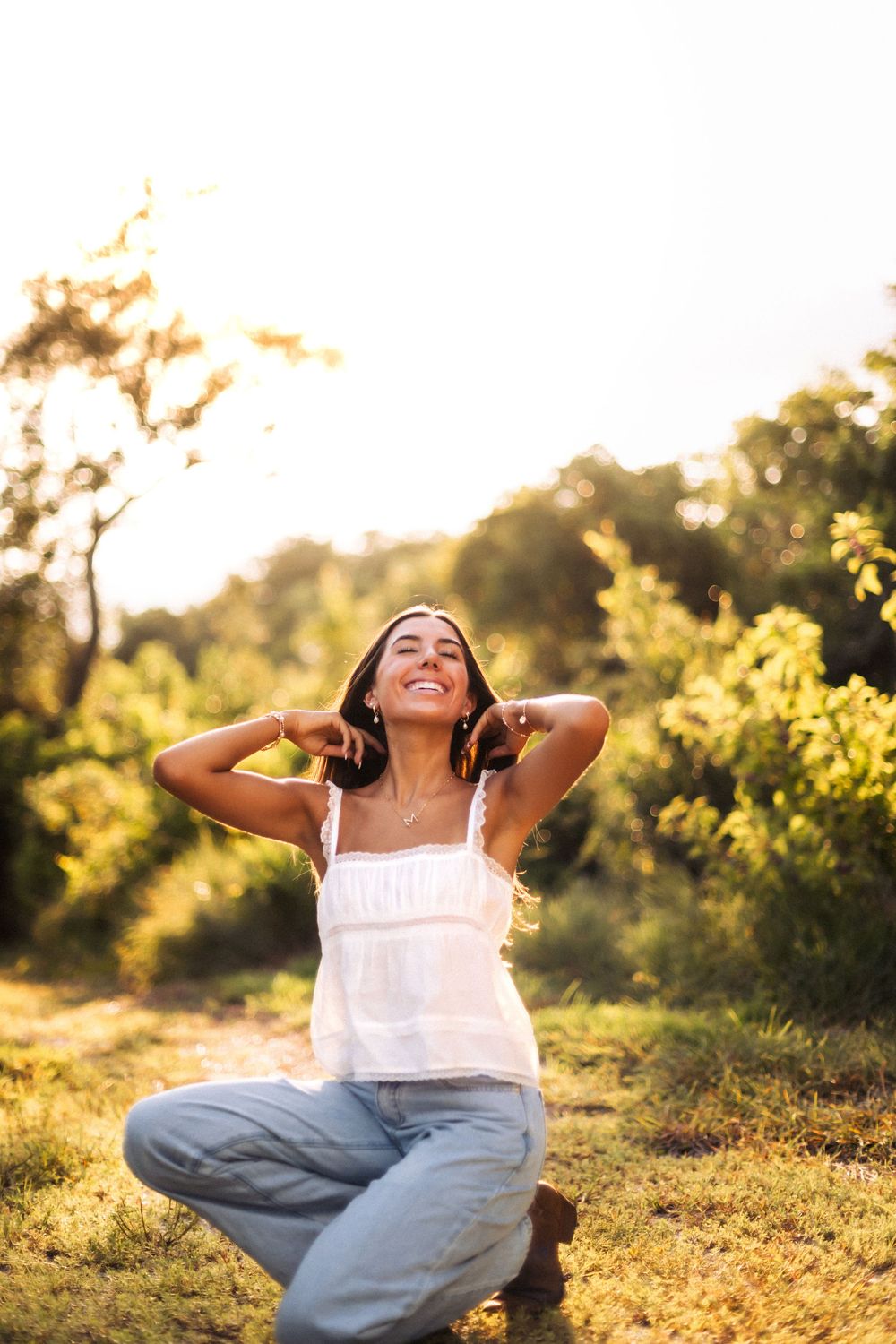 A smiling woman in a white top and jeans sits on grass in a sunlit park, hands behind her head.