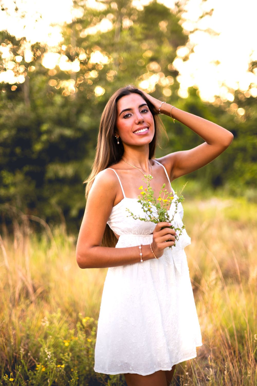 Young woman in white sundress holding wildflowers in a sunlit field, smiling with hand in hair.
