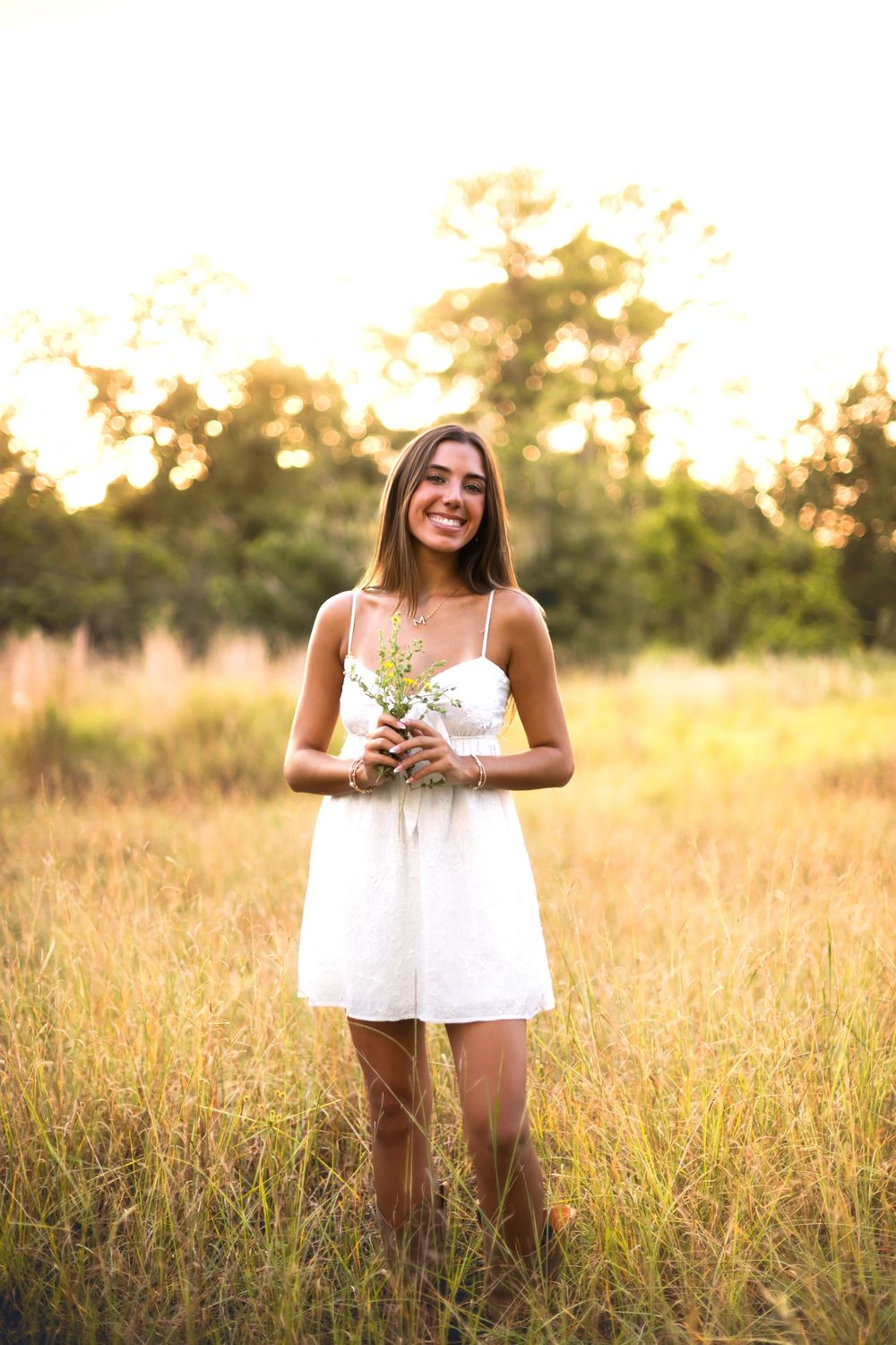 Young woman in white sundress holding wildflowers, smiling in golden hour sunlit meadow with trees in background.