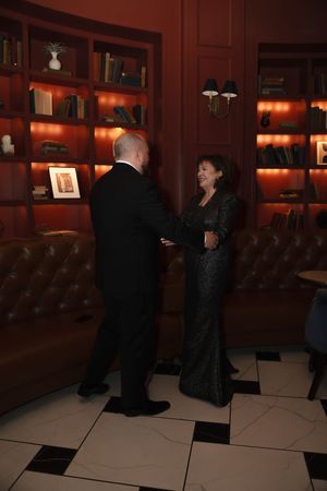 Elegant couple in formal attire dancing in a warmly lit library lounge with red bookshelves and checkered floor.