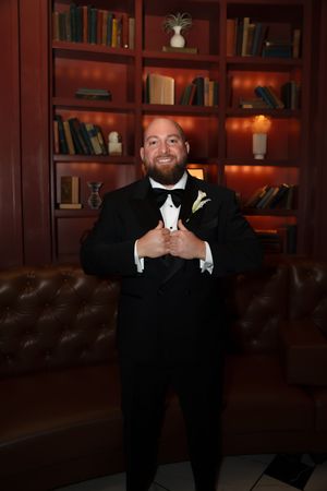 Smiling groom in black tuxedo with bow tie and boutonniere poses in front of wooden bookshelf.