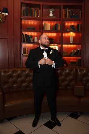 A groom in a black tuxedo with bow tie and boutonniere stands confidently before a warmly lit bookshelf backdrop.
