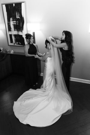 Two bridesmaids in black gowns help a bride with her long wedding veil in an elegant black and white photo.