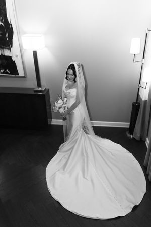 Black and white photo of a bride in a flowing white gown and veil, holding a bouquet in an elegant room.