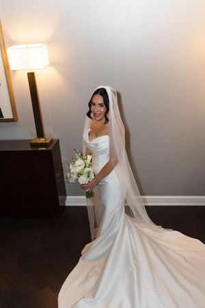 Elegant bride in a white satin mermaid gown with cathedral veil holding a white rose bouquet in a warmly lit room.