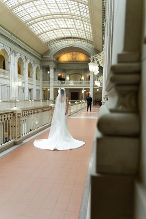 Bride in white gown with cathedral veil stands in grand neoclassical building with glass ceiling.