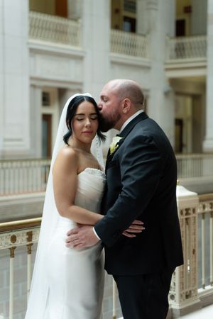 Groom kisses bride on the cheek as they embrace on a balcony in an elegant wedding venue.